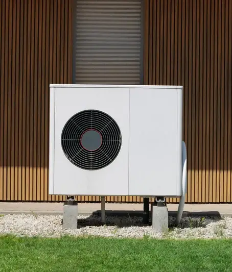 A large, white air conditioning unit is placed on concrete blocks outside a wooden slat building, surrounded by green grass and white gravel.