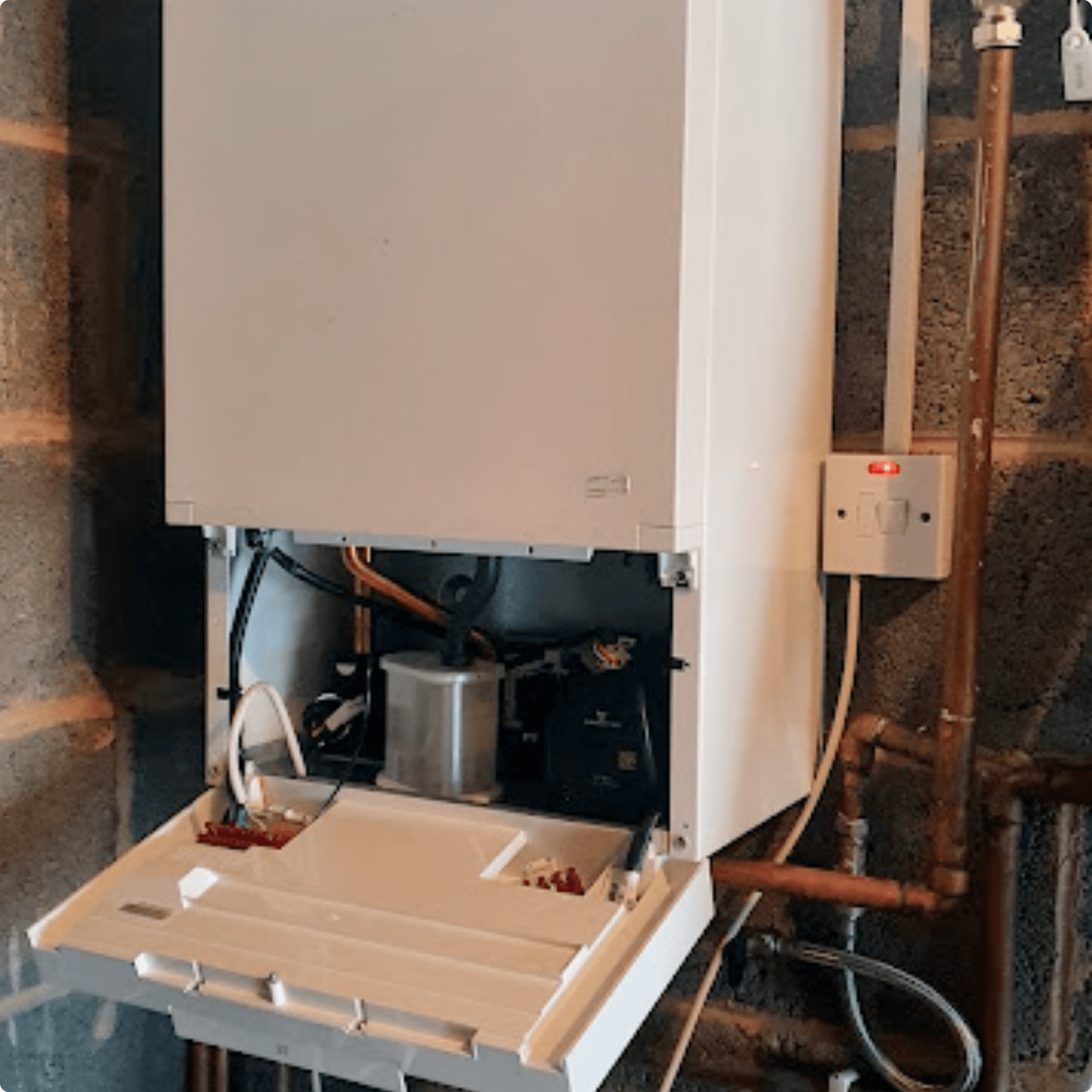 A white boiler with an open panel reveals internal components, mounted on a brick wall. Pipes and an electrical switch are visible nearby, conveying maintenance work.