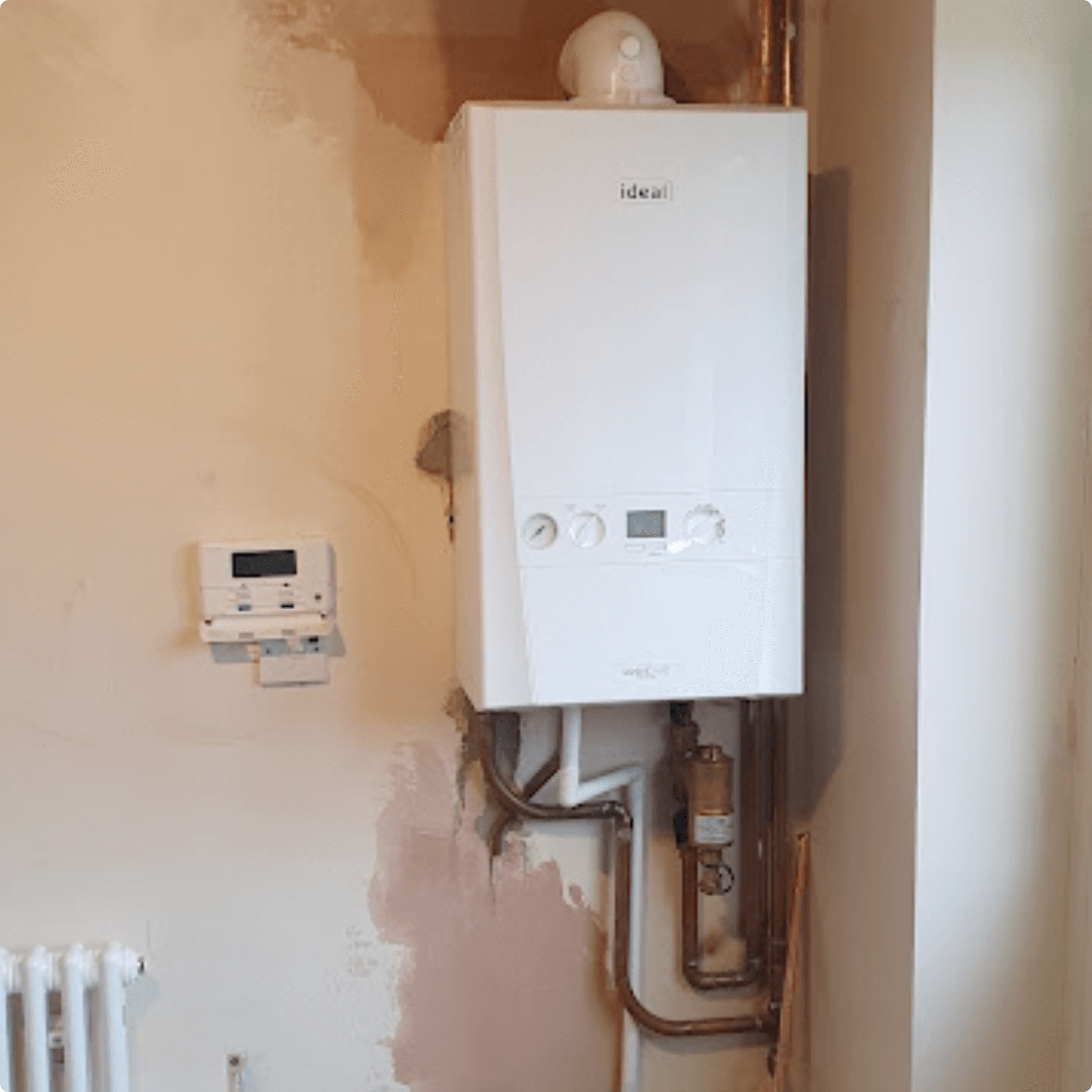 A white boiler mounted on a partially painted wall, with pipes beneath and a digital thermostat on the left. The setting is a utility room.