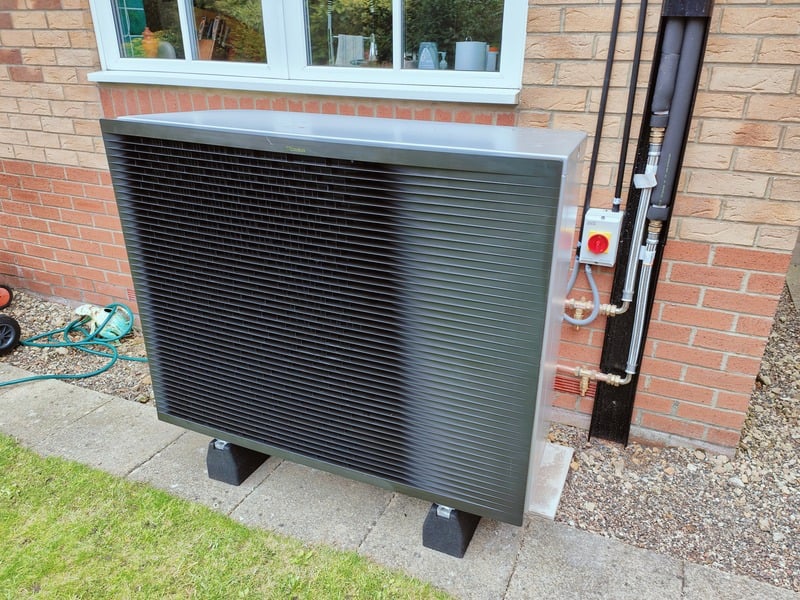 Outdoor heat pump unit mounted on a concrete slab next to a brick house, with connected pipes and a garden hose in the background.