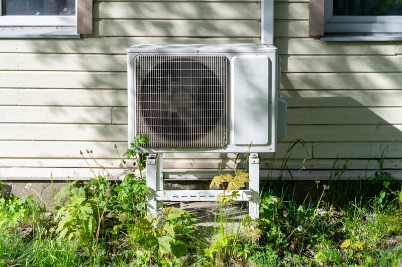 Outdoor air conditioning unit against a sunlit beige wooden wall, surrounded by overgrown green plants. The scene is calm and rustic.